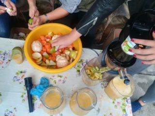 Na zdjęciu uczestnicy pikniku kroją warzywa i owoce i robią z nich sok za pomocą wyciskarki do owoców.
