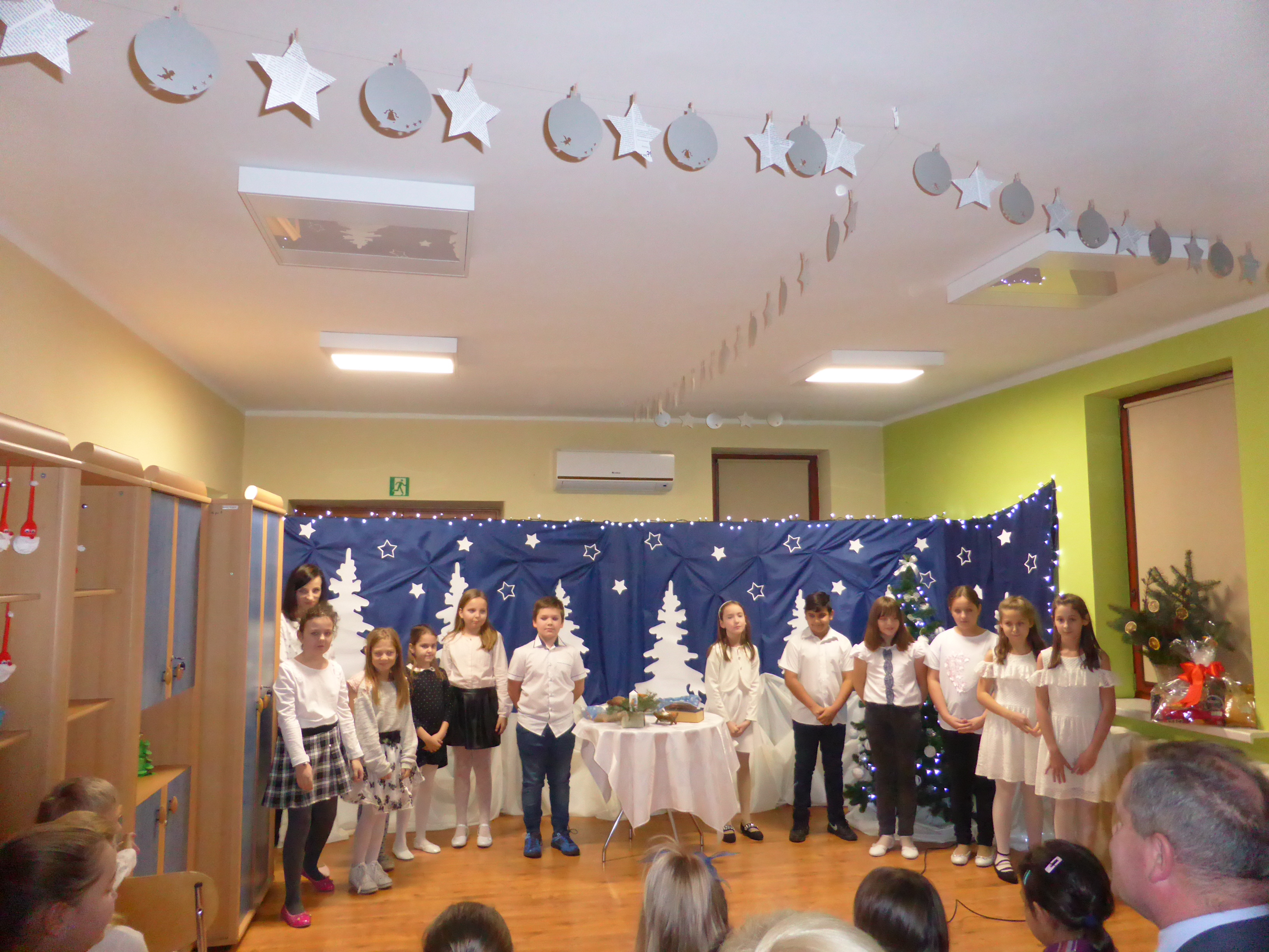 Na zdjęciu dzieci na scenie, za nimi dekoracja świąteczna - granatowy materiał, na nim papierowe choinki i gwiazdki