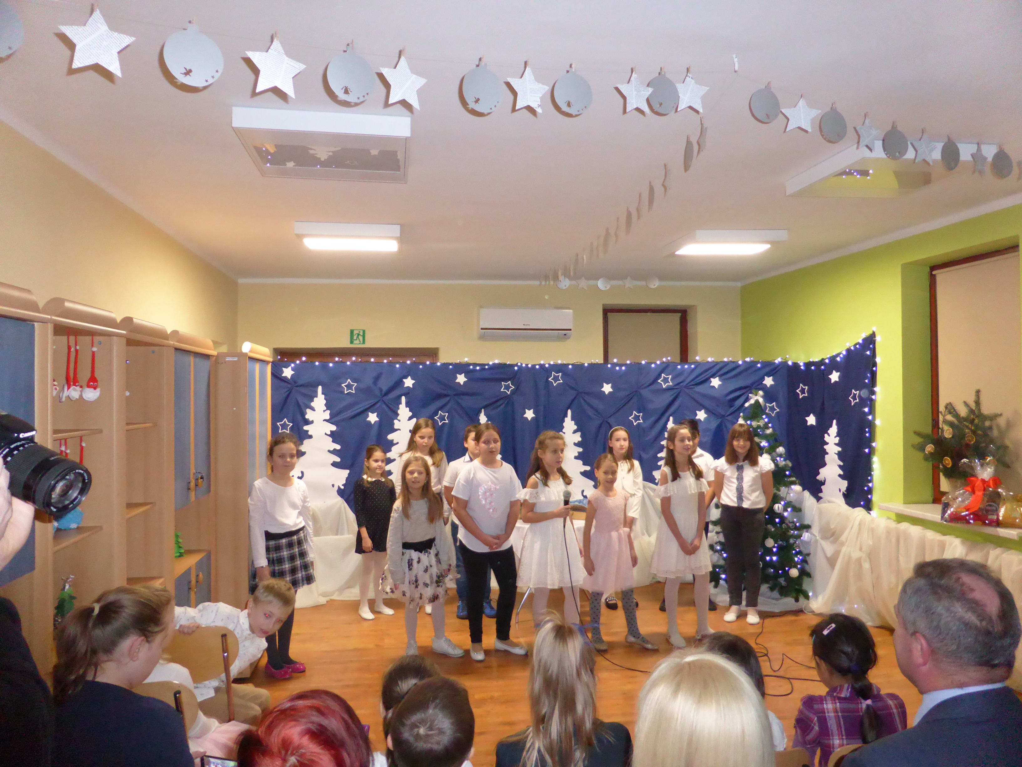 Na zdjęciu dzieci na scenie, za nimi dekoracja świąteczna - granatowy materiał, na nim papierowe choinki i gwiazdki