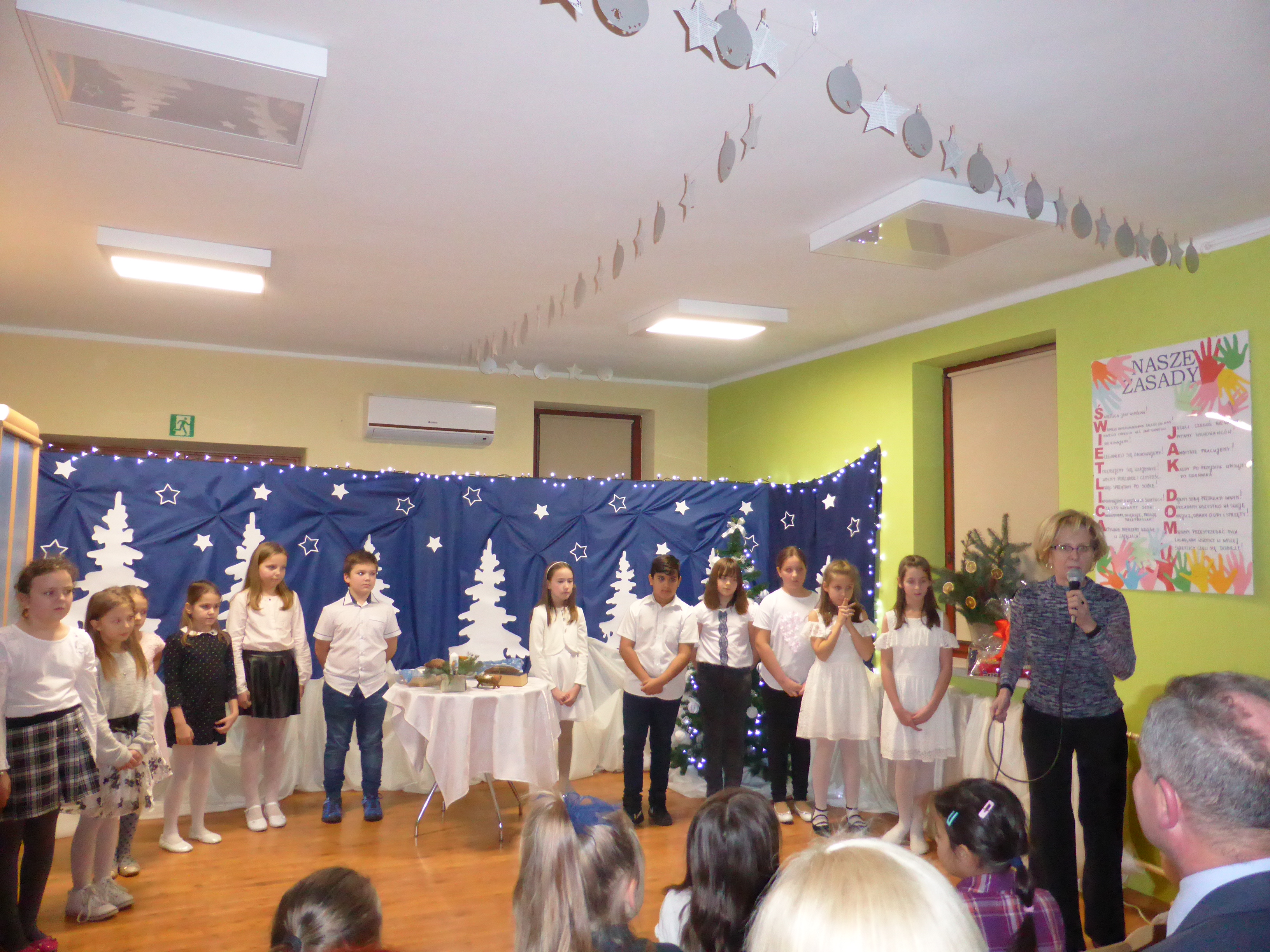Na zdjęciu dzieci na tle dekoracji, przed nimi przemawia dyrektor Ośrodka Pomocy Społecznej Elżbieta Prus