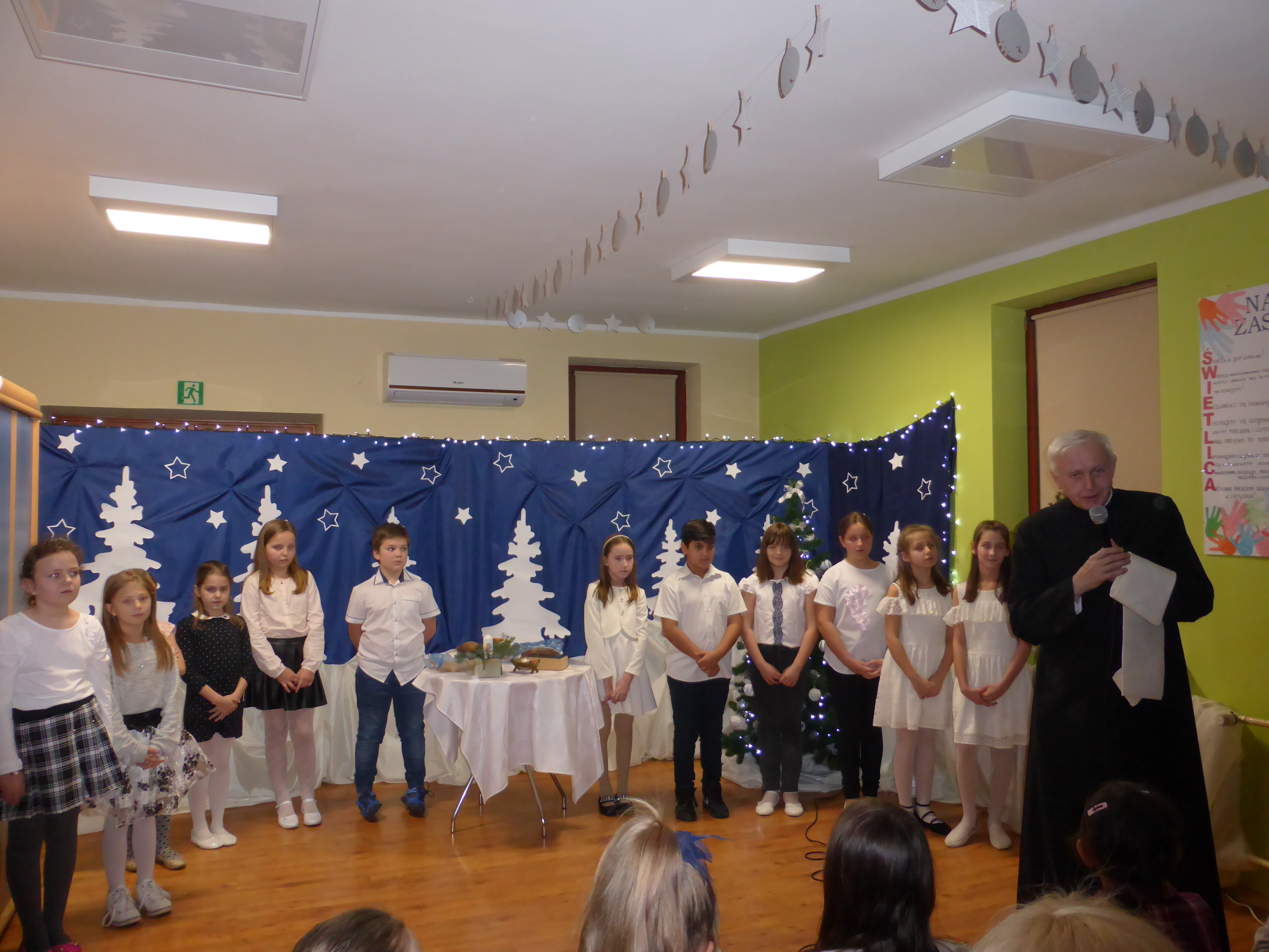 Na zdjęciu dzieci na tle dekoracji, przed nimi przemawia ksiądz prałat Stanisław Czernik, proboszcz parafii św. Macieja