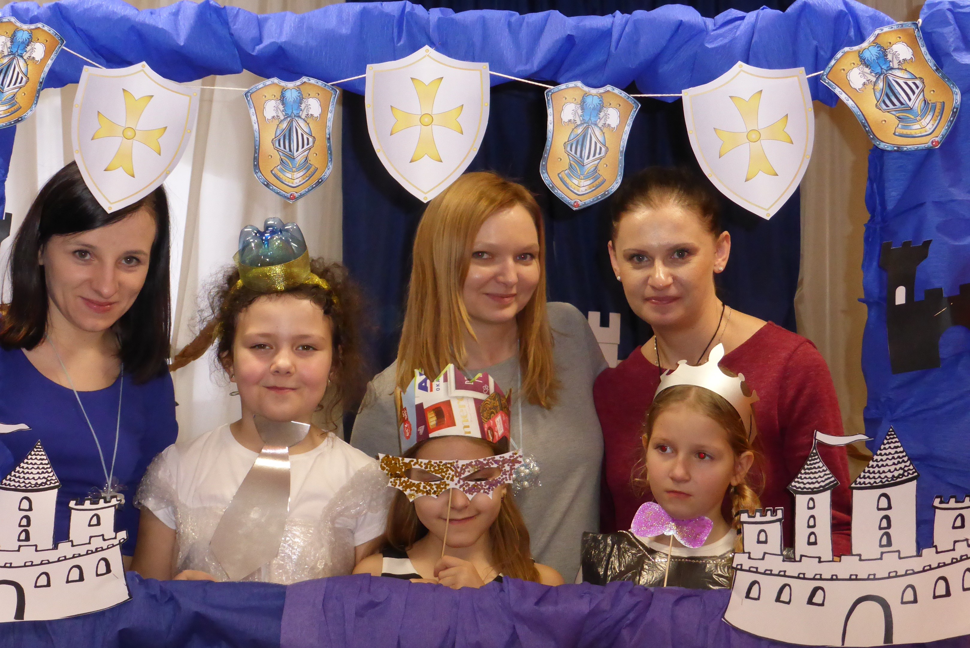 Na zdjęciu dziewczynki przebrane za księżniczki i panie wychowawczynie ze Świetlicy pozują do zdjęcia w ramce, do której przyczepione są papierowe tarcze z herbami