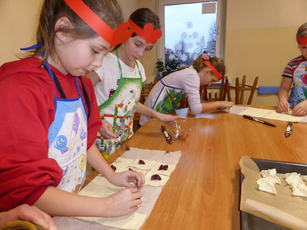 Na zdjęciu dzieci, które robią ciasteczka. Dzieci mają na głowach papierowe opaski z naklejonym sercem.