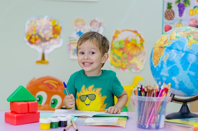 Zdjęcie uśmiechniętego chłopca, który siedzi przy biurku i pisze coś w zeszycie długopisem. Na biurku są jeszcze: globus, kredyt, klocki, farby i pędzle.