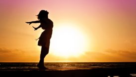 Kobieta stojąca na molo. W tle morze i zachodzące słońce. Kobieta wystawia twarz do wiatru, który rozwiewa jej włosy