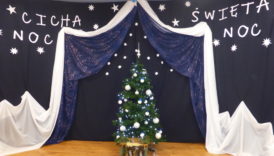 Scena ze świąteczną dekoracją - na granatowym tle białe ścieżynki z papieru i napis: Cicha noc, święta noc. Na środku sceny stoi choinka z białymi i niebieskimi bombkami, pod nią szopka.