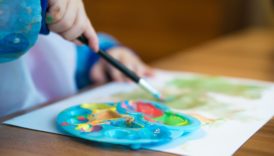 Dziecko maluje pędzlem na kartce papieru, obok paleta z farbami
