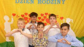 Chłopcy w odświętnych ubraniach składają dłonie w kształt serca, za nimi dekoracja - czerwone serca na żółto-zielonym tle i napis "Dzień Rodziny"