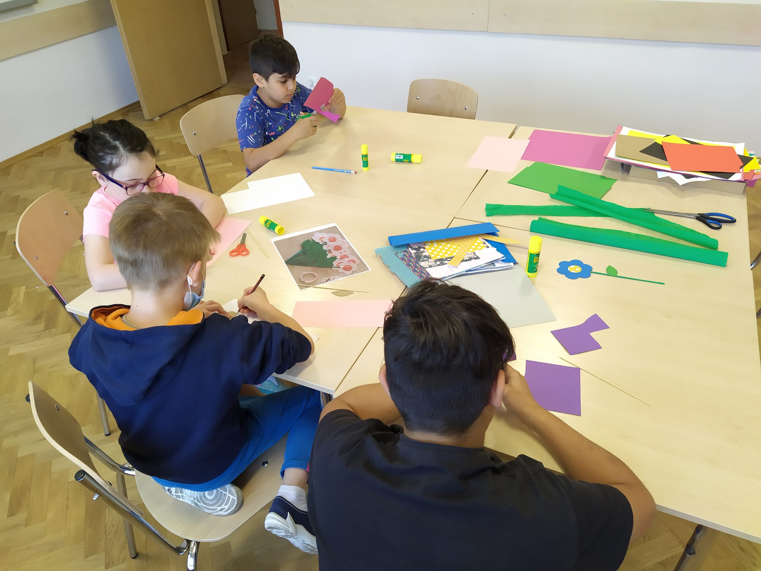 Dzieci siedzą wokół stołu, wycinają coś z papieru, na stole kolorowy papier, bibuła, nożyczki i kleje