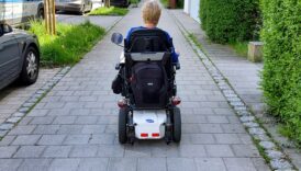 Mężczyzna jedzie na elektrycznym wózku inwalidzkim jedzie po chodniku, z prawej strony zabudowania, z lewej parking