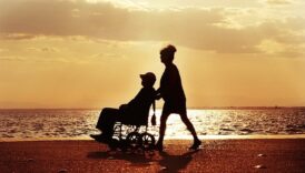 sylwetka kobiety pchającej wózek inwalidzki, na wózku sylwetka mężczyzny, w tle plaża, morze, zachodzące słońce