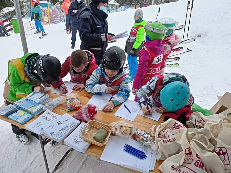 Dzieci gromadzą się wokół stołu, położonego na stoku narciarskim i rozwiązują krzyżówki