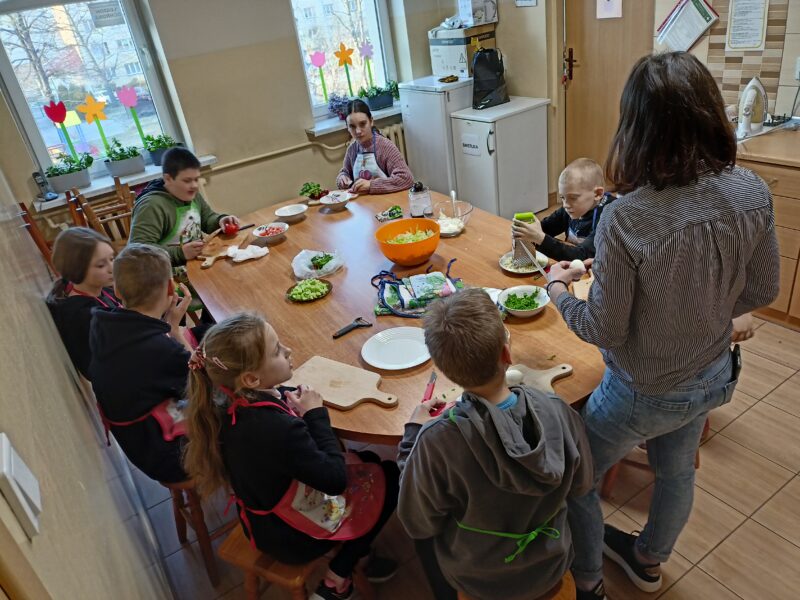 Grupa dzieci siedzi przy stole i przygotowują produkty na sałatkę warzywną