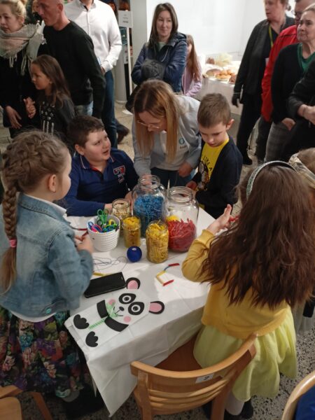 Dzieci siedzą przy stoliku i przygotowują naszyjniki z kolorowego makaronu