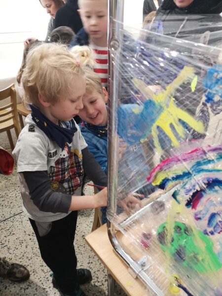 Dwójka dzieci maluje farbami po folii rozciągniętej na nogach od stołu