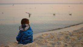 chłopiec w kurtce siedzi tyłem nad brzegiem morza