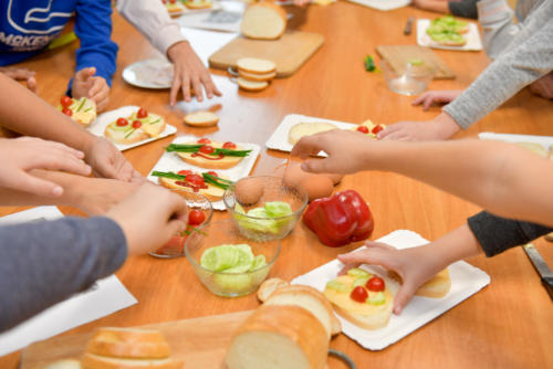 Ręce dzieci, które sięgają po produkty potrzebne do zrobienia kanapek, m.in. plasterki ogórka, pomidorki koktajlowe, paprykę i jajka.