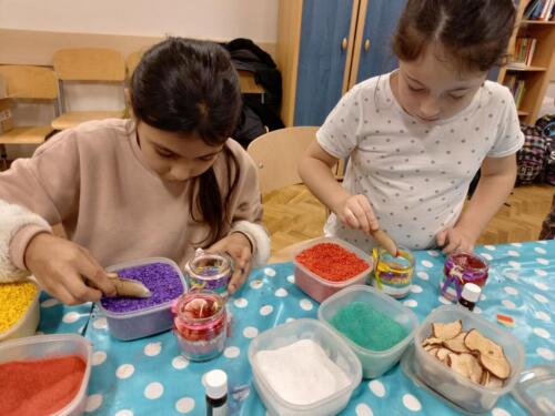 Dwie dziewczynki dekorują świeczki, wsypując kolorowe kamyczki do szklanych pojemników