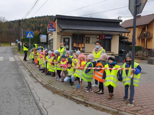 Małe dzieci stoją wraz z nauczycielami na chodniku przy drodze, mają kamizelki odblaskowe, obok strażnicy miejscy