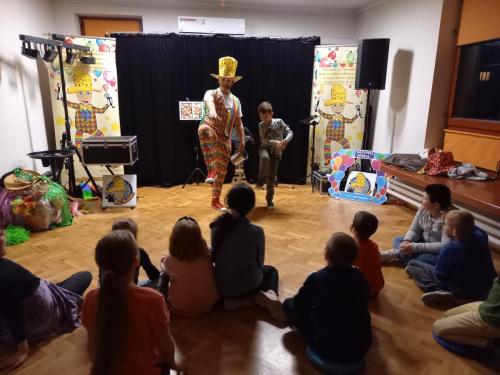 Pan Humorek wraz z chłopcem stoją na środku sali na jednej nodze, pozostałe dzieci oglądają występ, siedząc na podłodze.