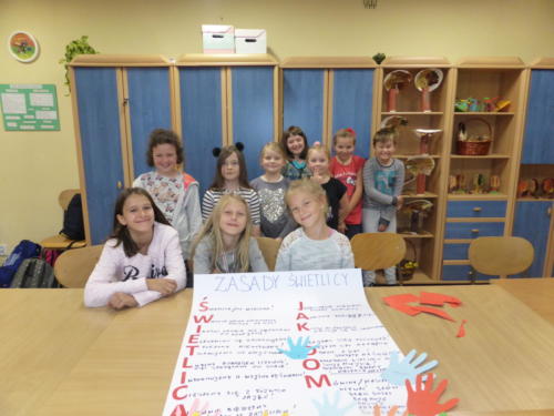Zdjęcie grupowe dzieci przy plakacie z zasadami świetlicy, ozdobionym odbiciami dłoni wyciętymi z papieru kolorowego